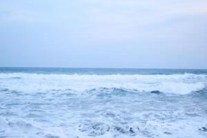 As the tide sweeps in at South Jogja Jetis Beach, Cornwall under a serene sky, waves gently roll, accompanied by ripples and bubbling foam. photo