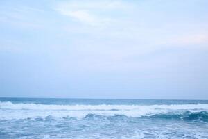 As the tide sweeps in at South Jogja Jetis Beach, Cornwall under a serene sky, waves gently roll, accompanied by ripples and bubbling foam. photo