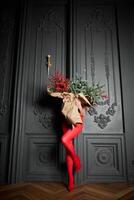un largo niña en rojo medias y tacones altos Zapatos sostiene un enorme ramo de flores de abeto ramas y rojo bayas envuelto en cartulina papel, ella cierra con un ramo, foto estudio con gris pared antecedentes