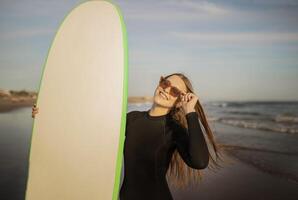 Portrait Of Beautiful Young Surfer Lady Holding Big Blank Surfboard, Smiling Millennial Woman Wearing Stylish Sunglasses And Wetsuit Standing On The Beach At Sunset, Enjoying Surfing, Copy Space photo