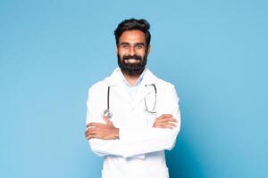Portrait of friendly indian male doctor in coat with stethoscope on neck posing with folded arms over blue studio background, looking and smiling at camera, free space photo