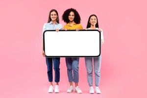 Full length shot of diverse ladies holding huge smartphone with white blank screen, offering mockup for your app or website design, pink studio background photo