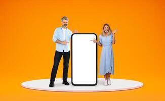 Happy beautiful middle aged spouses man and woman standing on platform by big phone with white empty screen, pointing at blank space, showing amazing online offer, deal, orange background photo