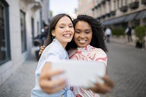 Two Beautiful Female Friends Laughing While Taking Selfie On Smartphone Outdoors, Happy Cheerful Multiethnic Besties Posing At Camera Together, Making Photos While Walking In City, Selective Focus