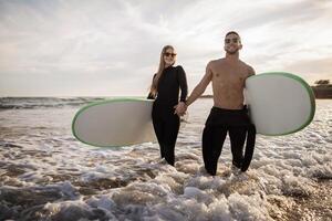 xtreme Deportes. joven deportivo Pareja surf juntos en el playa, retrato de sonriente hombre y mujer vistiendo trajes de neopreno participación manos y que lleva tablas de surf, milenario surfistas en pie en agua foto