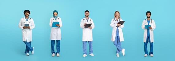 Smiling mature arabic and caucasian doctors in white coats with tablets and clipboards isolated on blue studio background, full length. Health care, exam, work, medicine and help photo