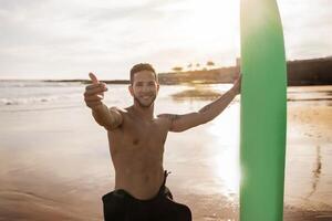 Surf With Me. Handsome Young Surfer Guy Holding Surfboard And Making Invite Gesture With Fingers At Camera, Shirtless Smiling Millennial Man Having Fun While Surfing On The Beach At Sunset photo