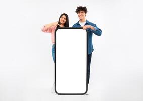 Great Mobile Application. Cheerful Young Couple Showing Huge Smartphone, Pointing Finger At Blank Screen For App Advertisement, Standing Behind Big Phone On White Studio Background. Mockup photo