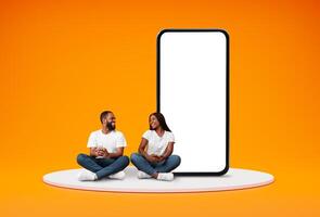 Happy Black Couple With Cellphones Sitting Near Bog Smartphone With Blank Screen, Cheerful African American Couple Browsing New Mobile App, Recommending Website, Posing On Orange Background, Mockup photo