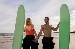 hermosa joven surfistas Pareja participación manos y posando con tablas de surf en el playa, contento romántico hombre y mujer vistiendo trajes de neopreno y Gafas de sol teniendo divertido juntos mientras surf, gratis espacio foto
