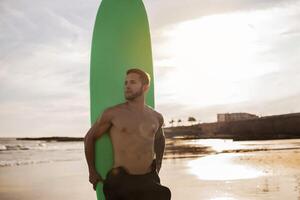Sporty Young Surfer Guy Posing With Surfboard On The Beach, Handsome Millennial Man Looking Aside, Athletic Shirtless Male Waiting For High Waves, Enjoying Surfing On Sunset Time, Copy Space photo