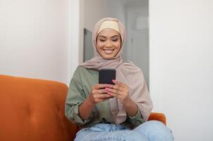 Mobile App. Happy Black Woman In Hijab Using Phone, Texting And Chatting In Social Media, Sitting On Couch At Home. Happy Islamic Lady Enjoying Gadget Use Browsing Internet On New Smartphone photo