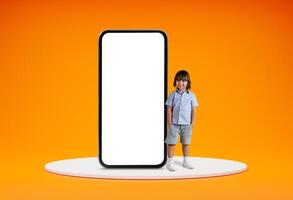 Entertaining mobile app for kids. Cute cheerful little boy pre-school age standing on platform by huge phone with white blank screen, orange studio background, blank space, mockup photo