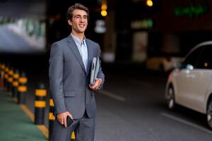 Handsome smiling millennial man wearing suit CEO manager standing at parking area, holding smartphone and laptop in his hands, waiting for his car or taxi, looking at copy space photo