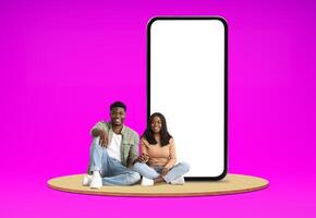 Relaxed positive happy millennial black man and woman lovers chilling on platform next to huge phone with white empty screen, showing nice dating mobile app, colored background, mockup, blank space photo