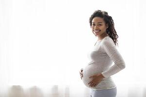 Happy black pregnant woman in comfortable clothes standing near window at home, smiling african american female embracing her big tummy, waiting for baby, enjoying pregnancy time, copy space photo