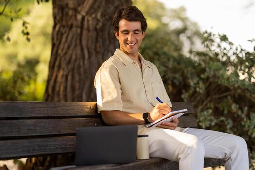 Cheerful young man attending webinar online course training while resting at park, guy sitting on bench at garden, looking at computer laptop screen, taking notes in notepad, learning new language photo