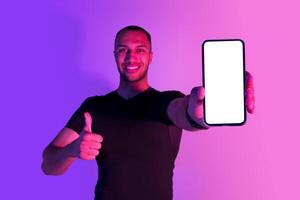 Happy black sportsman showing cellphone with empty screen and gesturing thumb up, smiling to camera, recommending workout and weight loss application, posing on purple neon background. Mockup photo