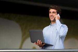 Communication in business concept. Friendly positive young entrepreneur with laptop computer in his hand have phone conversation with business partner or client, looking at copy space photo