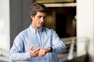 Serious handsome young businessman wearing formal outfit holding phone, standing at balcony in office building, checking his watch on wrist, getting ready before business meeting, copy space photo