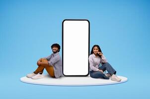 Cute cheerful smiling young eastern couple man and woman sitting on round platform by big phone with white blank screen, blue studio background, mockup blank space for mobile app, collage photo