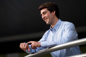 Young man employee using smart watch. Businessman uses smartwatch on his hand and keeps track of the time on the display while breathing fresh air next to office building, copy space photo