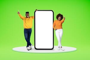 Dating mobile app concept. Happy millennial black man and woman in casual standing on platfotm by huge phone with white blank screen, showing thumb up, copy space, isolated on green background photo