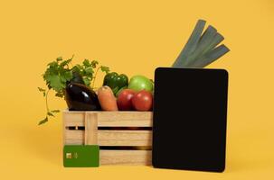 Online Grocery Shopping. Digital Tablet Computer With Empty Screen Standing Near Harvested Vegetables In Wooden Box On Yellow Studio Background. Mockup For Farm Business Advertisement photo
