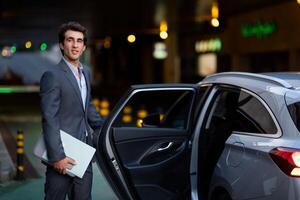 Attractive millennial man wearing expensive grey suit manager with laptop computer, phone and documents in his hand getting into luxury car in the evening, going home after working day photo