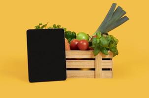 Digital Tablet With Blank Screen Standing Near Wooden Box With Fresh Vegetables And Fruits Over Yellow Studio Background. Mockup For Agriculture Website Advertisement Or Online Grocery Shopping photo