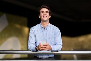 Happy handsome young guy businessman entrepreneur searching information on smartphone and smiling. Millennial caucasian man manager using newest business mobile app, standing at office building photo
