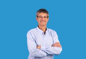 Confident business person. Happy mature businessman in shirt posing and crossing hands with expression of confidence, smiling looking at camera standing over blue background, studio shot photo