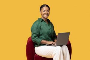Website Ad. Smiling African American Woman Using Laptop Computer Sitting In Red Armchair Working Online And Surfing Internet Over Yellow Background, Studio Shot. Freelancer Posing With PC on Lap photo