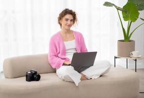 Portrait Of Smiling Young Woman With Laptop Sitting On Couch Next To Photocamera, Beautiful Millennial Freelance Photographer Lady Using Computer At Home For Editing Photos, Copy Space photo