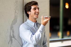 Handsome young man in fromal outfit employee standing at parking place next to office building, using smartphone and wireless earphones outdoor, recording voicemail, chatting, copy space photo