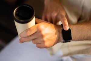 Close up shot of a businessman hand that uses a smart watch to view incoming messages and calls while sitting outdoors and drinking takeaway coffee, the gadget simplifies his life, cropped photo