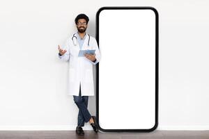 Telehealth app. Positive handsome millennial eastern doctor holding medical chart wearing white medical coat standing by huge smartphone with white empty screen mockup, isolated on white background photo