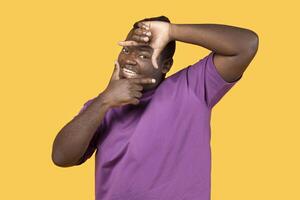 Capturing moments. Happy black man making picture frame with fingers, looking at camera and smiling for photo posing over yellow studio background. African american guy showing photograph gesture