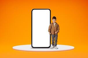 Educational kids mobile app. Happy positive cute little boy standing by big smartphone with black white screen and smiling, ready for school, posing over orange studio background, copy space photo