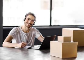 New Job. Happy Young Manager Guy With Headset Taking Notes At Laptop, While Communicating With Internet Clients, Enjoying First Day At Workplace, Sitting At Table With Boxes In Office Indoor photo