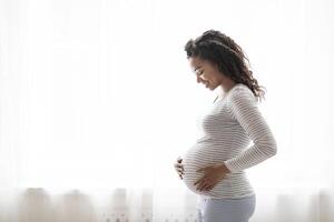 Beautiful pregnant black woman hugging her tummy while standing near window at home, happy african american female enjoying her pregnancy, touching big belly and smiling, side view with copy space photo