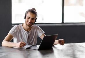E-Learning. Young Middle Eastern Guy Taking Notes Learning At Laptop, Smiling Posing With Headset Sitting At Desk In Office Interior, Free Space. Man Studying And Working Online At Workplace photo