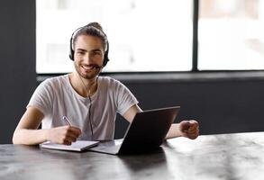 Smiling Hispanic Young Man Sitting At Laptop Computer Wearing Headset While Working, Communicating And Learning Via Online Call Sitting At Desk In Modern Office, Looking At Camera photo