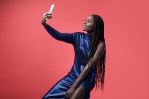 Smiling African American Woman In Sequin Dress Taking Selfie On Smartphone While Sitting On Chair Over Red Studio Background, Stylish Young Black Female Making Self-Portrait, Posing At Mobile Camera photo