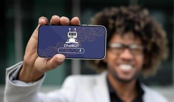Positive handsome young african american guy entrepreneur showing horizontal phone with chatbot application on screen. Businessman using artificial intelligence in business process, collage photo