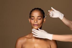 Cosmetic beauty injections concept. Attractive black middle aged woman getting facial injection in cheekbones zone, standing over brown studio background, free space photo