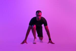 Front View Of Motivated African American Runner Guy Doing Crouch Start And Looking At Camera, Sportsman Posing Ready For Race On Purple Neon Background. Fit Man Preparing For Running Workout photo