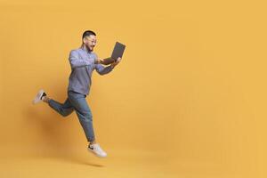 Online Sales. Excited Young Asian Man With Laptop Computer Running Over Yellow Background, Happy Millennial Male Looking At Screen And Smiling, Enjoying Website Offer, Full Length, Copy Space photo