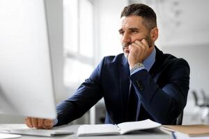 Bored middle aged businessman sitting in front of computer at workplace in office, leaning head on hand and looking at screen. Upset man suffering job problems, reading bad negative news photo