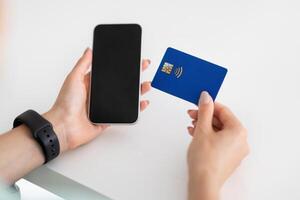 Hands of young european woman use smartphone with blank screen and credit card on white table in office, cropped. Banking, business and work, device and shopping, sale app, order and pay photo